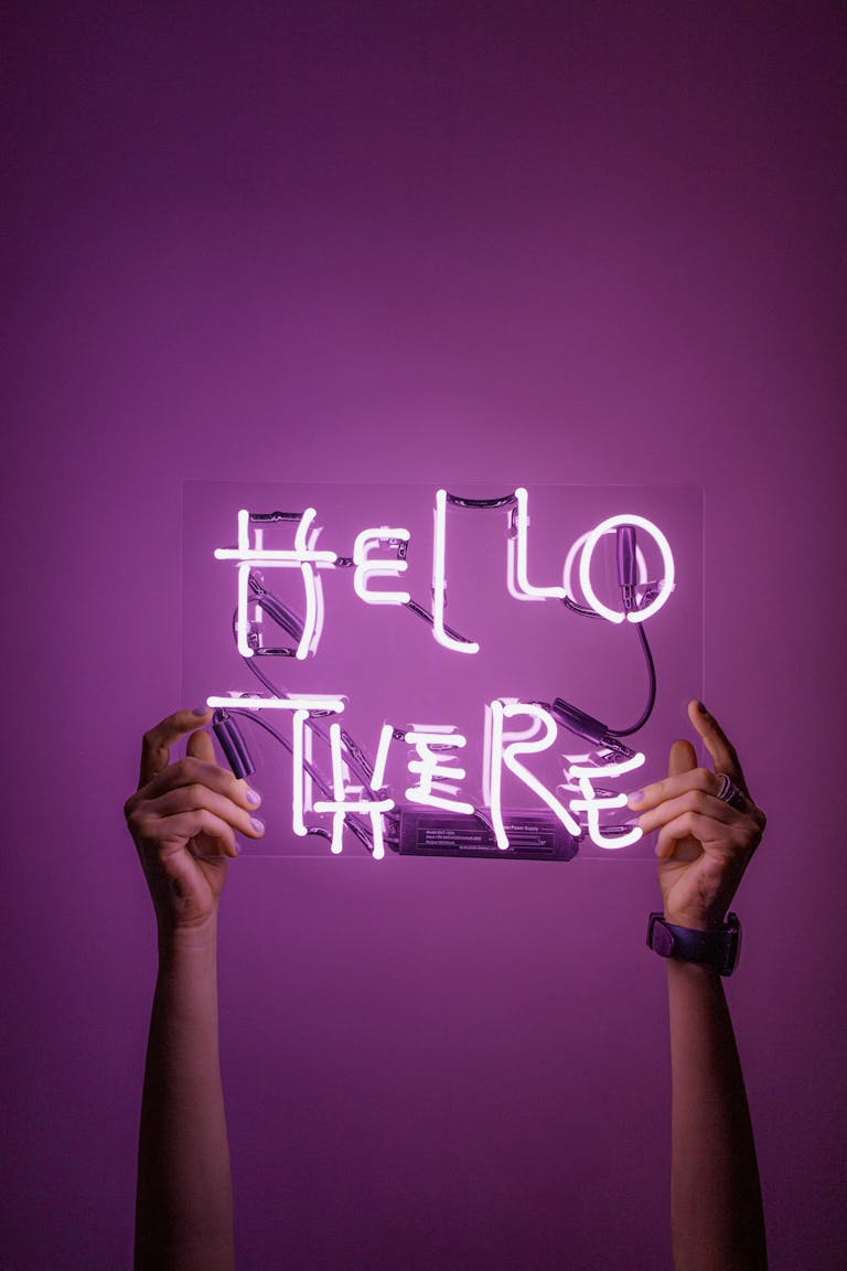 Bright neon sign saying 'Hello There' held by hands against a purple background.